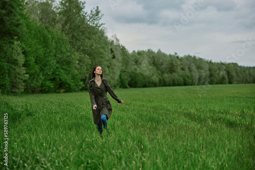 a beautiful woman in a long raincoat runs across a field in high grass in spring in cloudy weather © VICHIZH