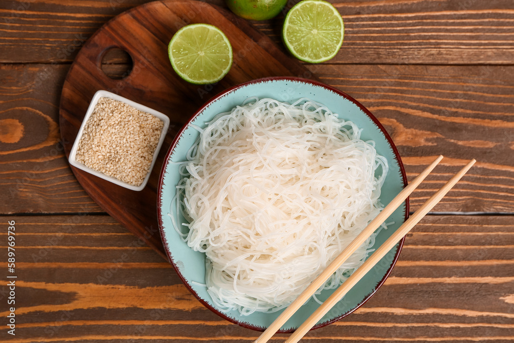 Bowl with tasty rice noodles, chopsticks, sesame seeds and lime on brown wooden table Bowl with tasty rice noodles, chopsticks, sesame seeds and lime on brown wooden table