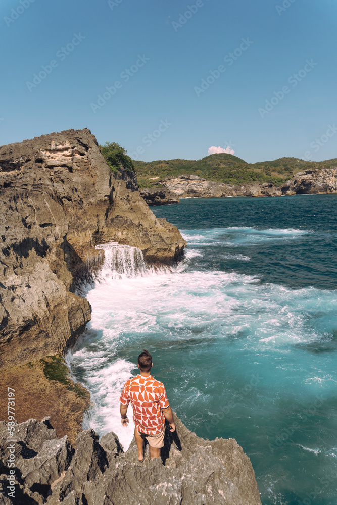 Podróżnik stojący na klifie, na brzegu skał, na tle fal i oceanu. - obrazy, fototapety, plakaty 