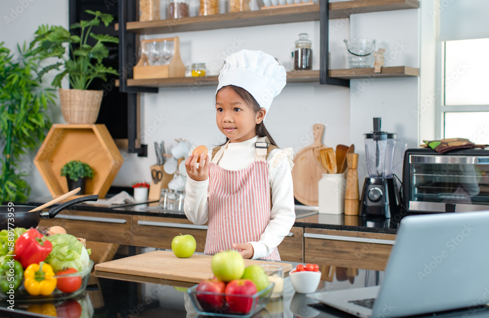 Millennial Asian young little cute girl chef daughter with white tall cook hat and apron standing smiling holding wooden spoon and fork posing ready for cooking vegetables at counter in home kitchen Millennial Asian young little cute girl chef daughter with white tall cook hat and apron standing smiling holding wooden spoon and fork posing ready for cooking vegetables at counter in home kitchen