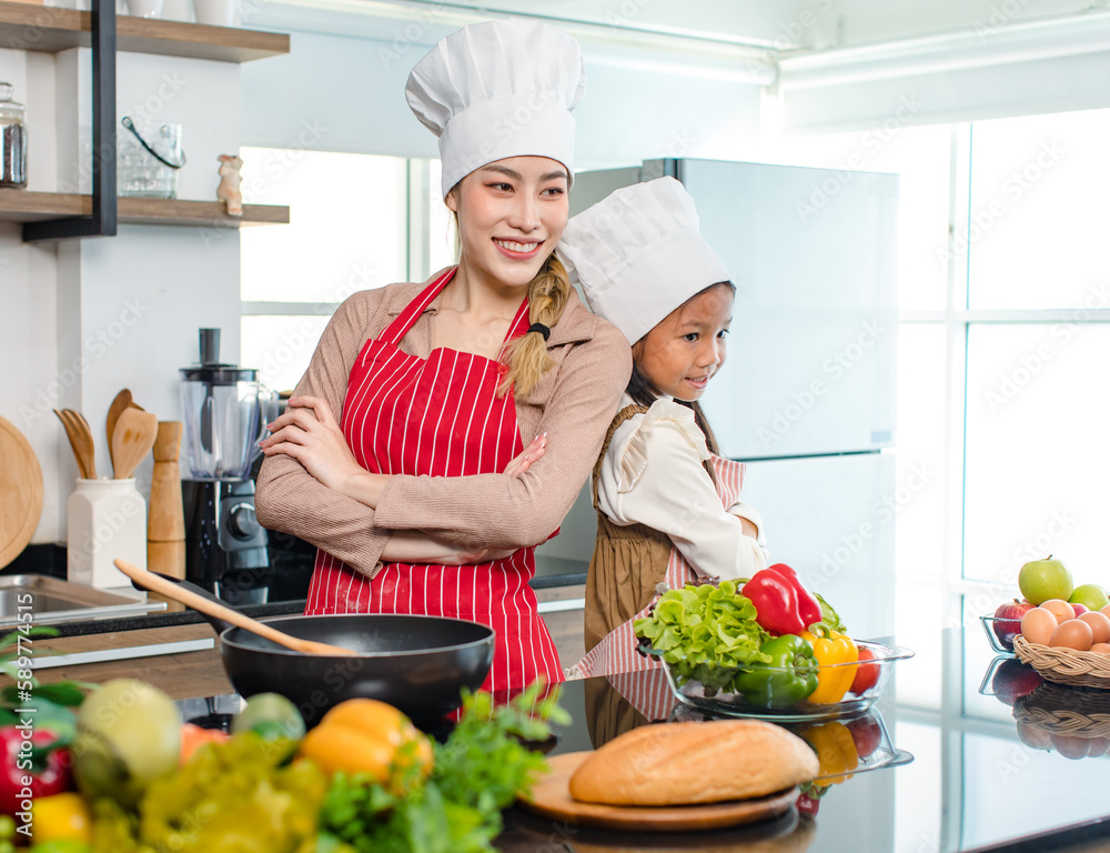 Asian young cheerful female mother and little cute girl daughter wearing white tall cook hat and apron standing crossed arms smiling posing holding thumbs up together ready for cooking in kitchen Asian young cheerful female mother and little cute girl daughter wearing white tall cook hat and apron standing crossed arms smiling posing holding thumbs up together ready for cooking in kitchen