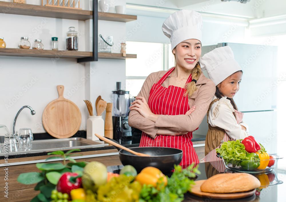 Asian young cheerful female mother and little cute girl daughter wearing white tall cook hat and apron standing crossed arms smiling posing holding thumbs up together ready for cooking in kitchen Asian young cheerful female mother and little cute girl daughter wearing white tall cook hat and apron standing crossed arms smiling posing holding thumbs up together ready for cooking in kitchen