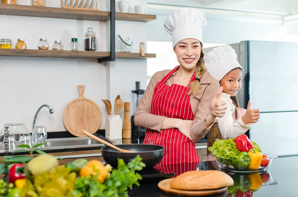 Asian young cheerful female mother and little cute girl daughter wearing white tall cook hat and apron standing crossed arms smiling posing holding thumbs up together ready for cooking in kitchen Asian young cheerful female mother and little cute girl daughter wearing white tall cook hat and apron standing crossed arms smiling posing holding thumbs up together ready for cooking in kitchen