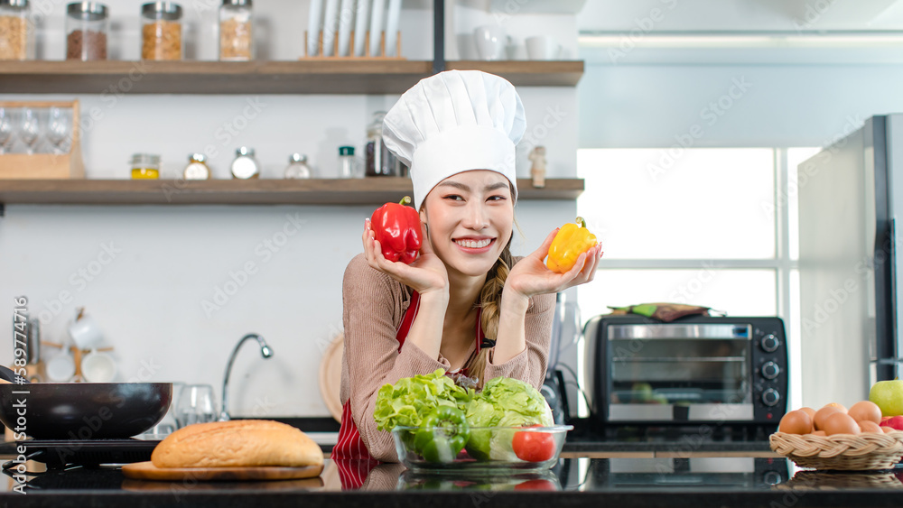 Asian young female chef wears white tall cook hat and apron smiling posing holding green apple ready to cooking food with pan at counter with bread vegetables ingredients in home kitchen.