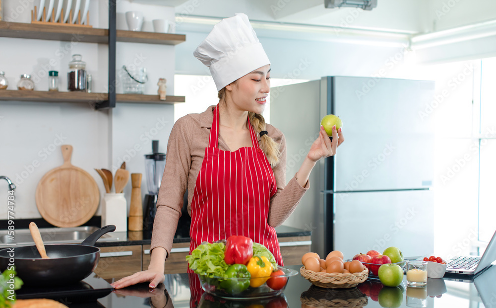 Asian young female chef wears white tall cook hat and apron smiling posing holding green apple ready to cooking food with pan at counter with bread vegetables ingredients in home kitchen. Asian young female chef wears white tall cook hat and apron smiling posing holding green apple ready to cooking food with pan at counter with bread vegetables ingredients in home kitchen.