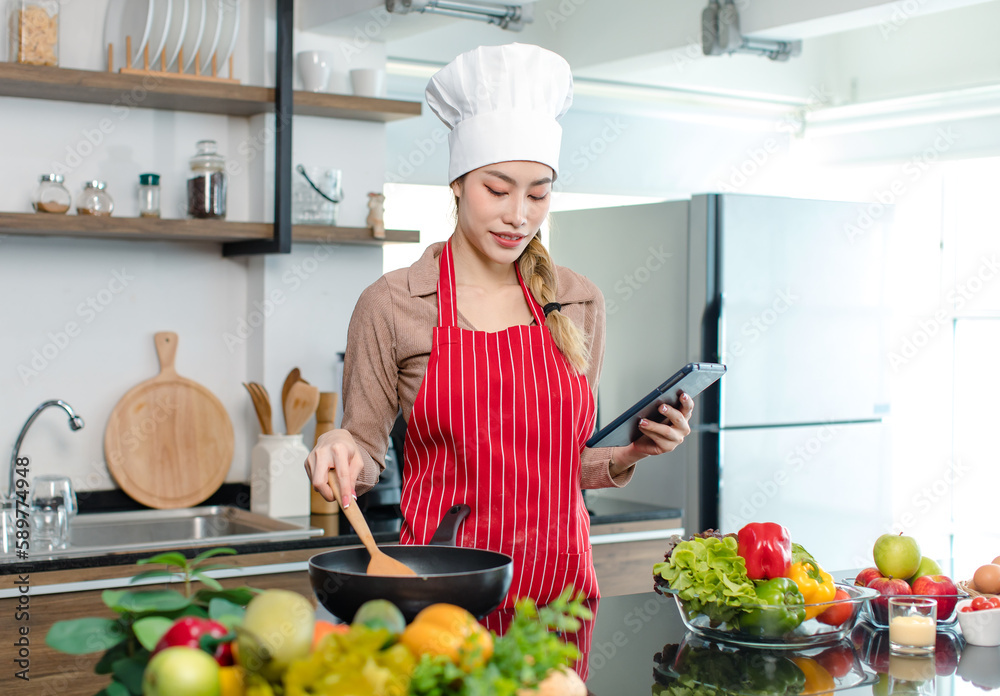 Asian young female chef housewife wears white tall cook hat and apron learning cooking homemade food meal online via touchscreen tablet computer while using spatula frying eggs in pan at home kitchen Asian young female chef housewife wears white tall cook hat and apron learning cooking homemade food meal online via touchscreen tablet computer while using spatula frying eggs in pan at home kitchen
