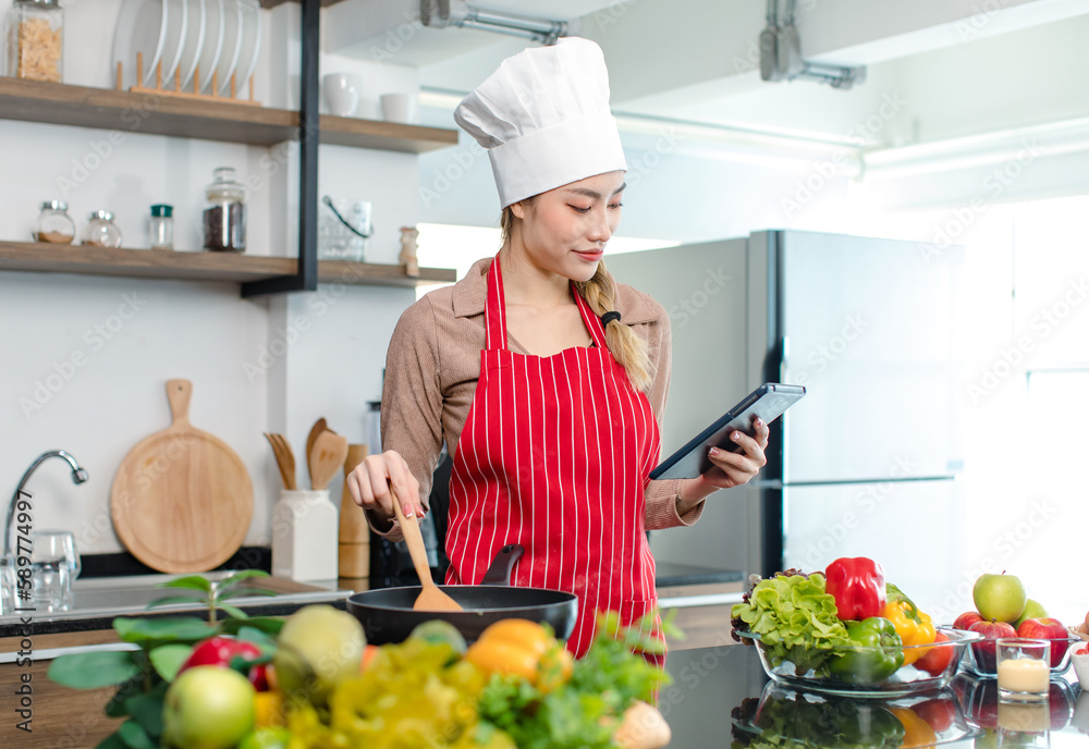 Asian young female chef housewife wears white tall cook hat and apron learning cooking homemade food meal online via touchscreen tablet computer while using spatula frying eggs in pan at home kitchen Asian young female chef housewife wears white tall cook hat and apron learning cooking homemade food meal online via touchscreen tablet computer while using spatula frying eggs in pan at home kitchen