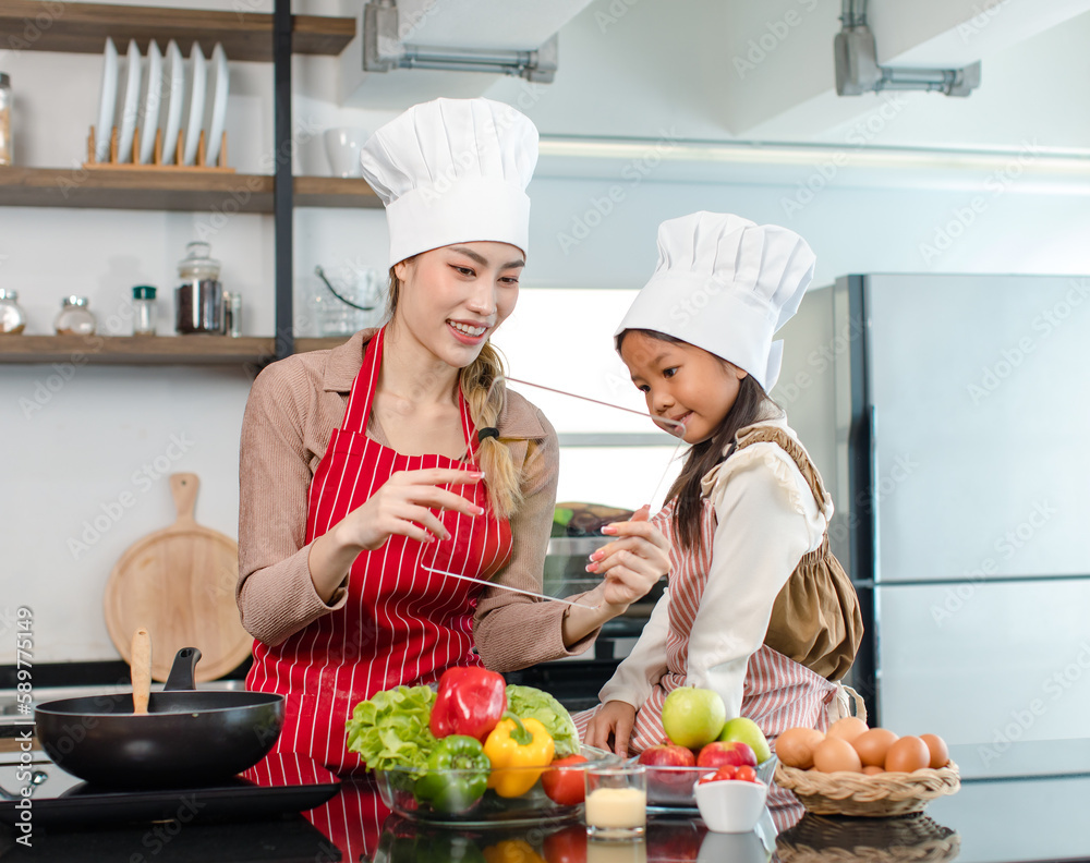 Asian young female chef housewife mother wears white tall cook hat and apron showing mockup glass touchscreen tablet computer screen to little cute girl chef learning cooking online at home kitchen Asian young female chef housewife mother wears white tall cook hat and apron showing mockup glass touchscreen tablet computer screen to little cute girl chef learning cooking online at home kitchen