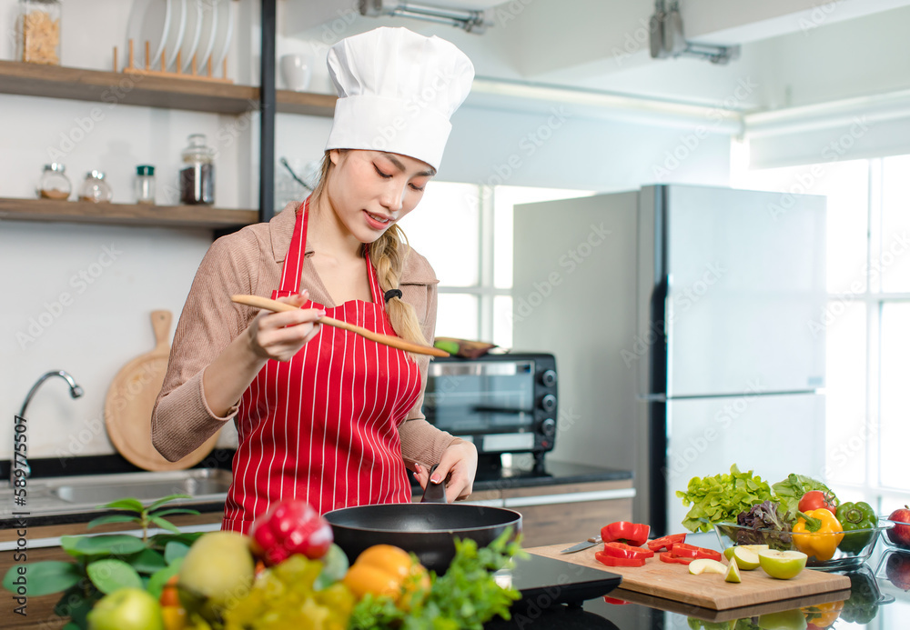 Asian young female chef housewife wears white tall cook hat and apron standing holding wooden spatula spoon tasting food meal from frying pan while cooking at counter with vegetables in home kitchen Asian young female chef housewife wears white tall cook hat and apron standing holding wooden spatula spoon tasting food meal from frying pan while cooking at counter with vegetables in home kitchen