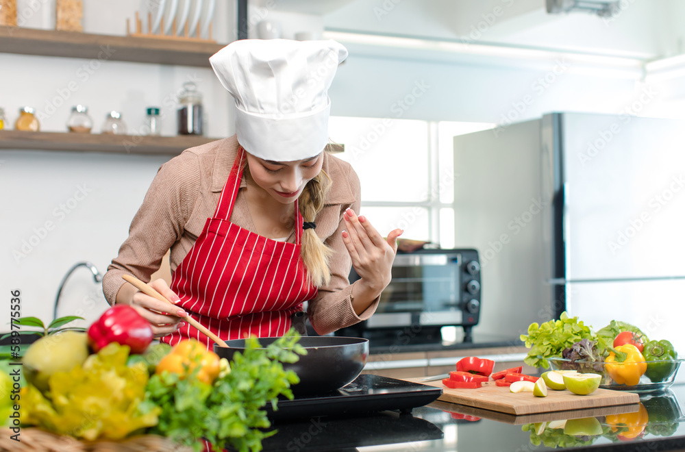 Millennial Asian young cheerful female housewife chef wearing white tall cook hat and apron standing smiling waving hand smelling tasty delicious meal while cooking in pan in full decorated kitchen Millennial Asian young cheerful female housewife chef wearing white tall cook hat and apron standing smiling waving hand smelling tasty delicious meal while cooking in pan in full decorated kitchen