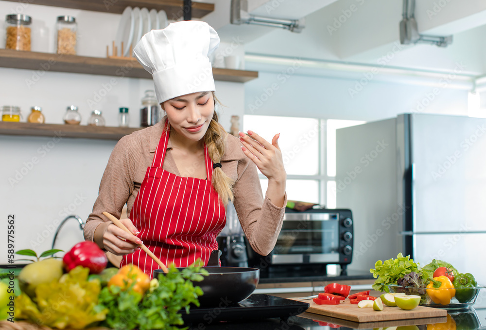 Millennial Asian young cheerful female housewife chef wearing white tall cook hat and apron standing smiling waving hand smelling tasty delicious meal while cooking in pan in full decorated kitchen Millennial Asian young cheerful female housewife chef wearing white tall cook hat and apron standing smiling waving hand smelling tasty delicious meal while cooking in pan in full decorated kitchen