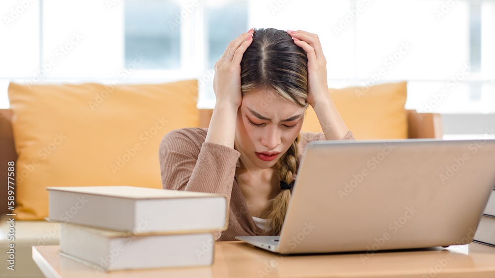 Asian upset unhappy stressed frowning face young female college student sitting working studying learning via laptop computer and textbooks in living room at home preparing for examination at campus