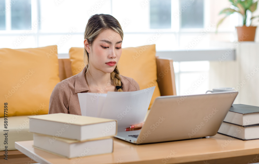 Asian beautiful young female college student sitting working studying learning via laptop computer and textbooks in living room at home preparing for examination at campus Asian beautiful young female college student sitting working studying learning via laptop computer and textbooks in living room at home preparing for examination at campus