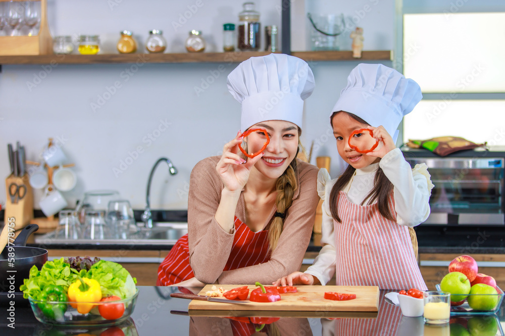 Asian young female chef housewife mother and little cute girl daughter wears white tall cook hat and apron smiling holding posing looking through sliced red sweet pepper with knife at home kitchen Asian young female chef housewife mother and little cute girl daughter wears white tall cook hat and apron smiling holding posing looking through sliced red sweet pepper with knife at home kitchen