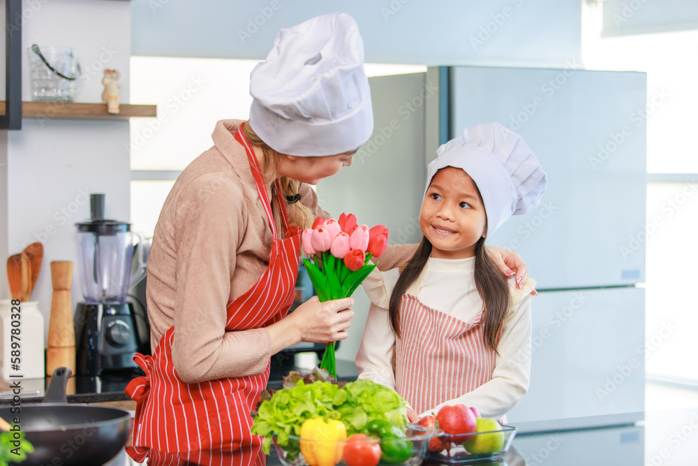 Asian young cheerful female chef housewife mother and little girl daughter wears white tall cook hat and apron standing smiling holding tulip flower bouquet together in kitchen preparing food meal Asian young cheerful female chef housewife mother and little girl daughter wears white tall cook hat and apron standing smiling holding tulip flower bouquet together in kitchen preparing food meal