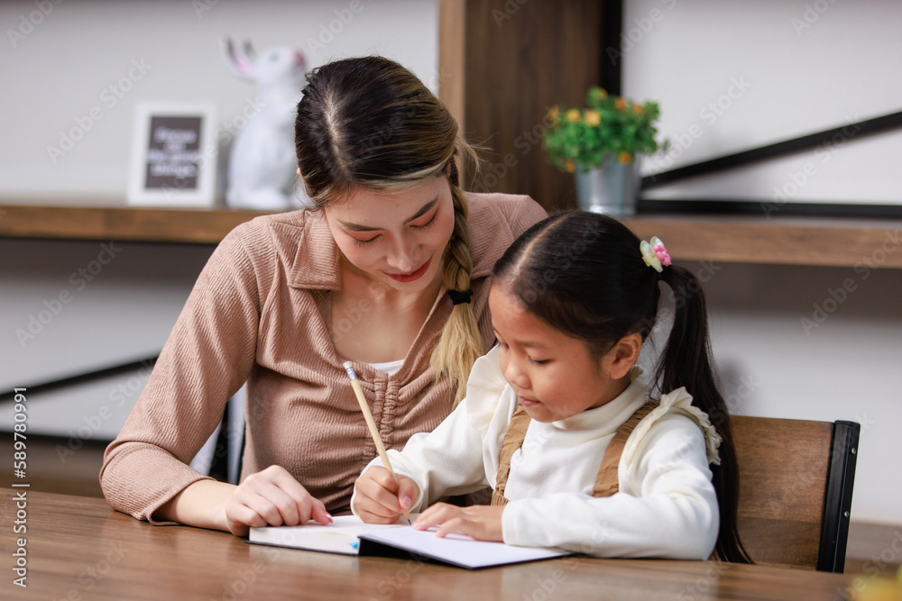 Asian young female housewife mother tutor teacher sitting smiling on table in living room at home teaching little cute kindergarten preschool girl daughter writing on book doing homework after school Asian young female housewife mother tutor teacher sitting smiling on table in living room at home teaching little cute kindergarten preschool girl daughter writing on book doing homework after school