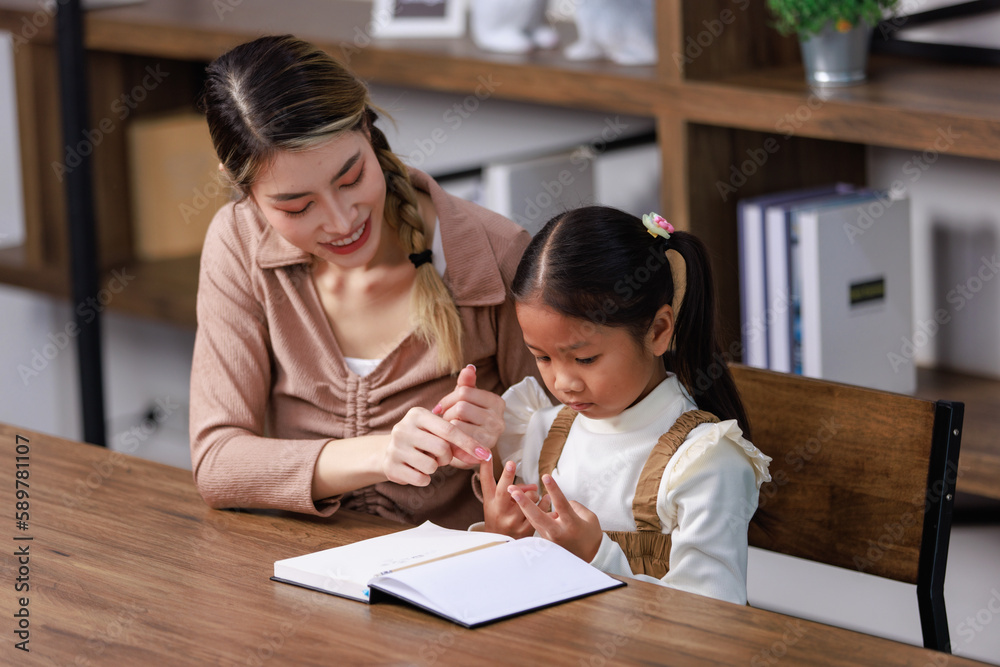 Asian young female housewife mother tutor teacher sitting smiling on table in living room at home teaching little cute kindergarten preschool girl daughter writing on book doing homework after school Asian young female housewife mother tutor teacher sitting smiling on table in living room at home teaching little cute kindergarten preschool girl daughter writing on book doing homework after school