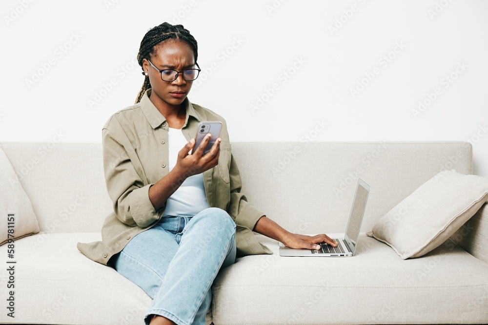 African American woman business freelancer working sitting on the couch at home in a laptop and phone, business calls and correspondence sadness and anger, home clothes and eyeglasses, light interior African American woman business freelancer working sitting on the couch at home in a laptop and phone, business calls and correspondence sadness and anger, home clothes and eyeglasses, light interior