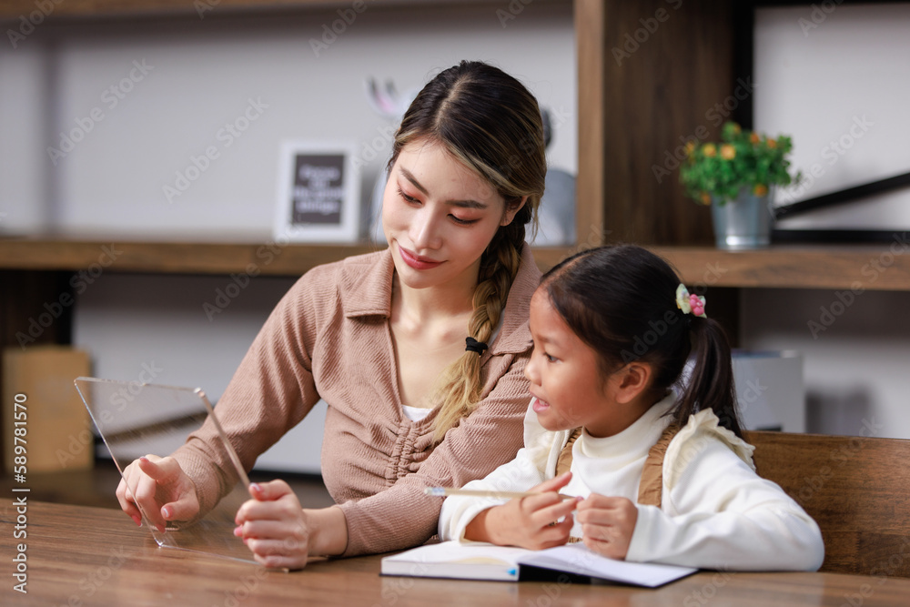 Fototapeta premium Asian young female housewife mother tutor teacher sitting smiling on table in living room holding tablet computer pointing teaching little cute kindergarten preschool girl daughter doing homework