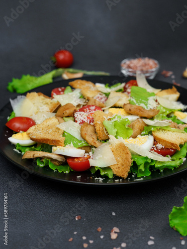 Close-up of a Caesar Salad with chicken, lettuce leaves, cherry tomatoes, grated parmesan in a black plate against a Black Background
