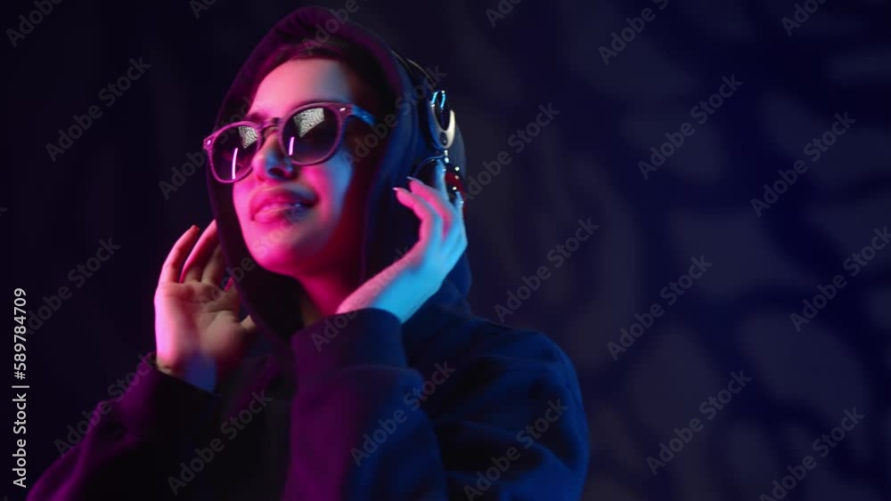 Fashion woman in hood sunglasses listening music in headphones clubbing at neon ultraviolet backlit