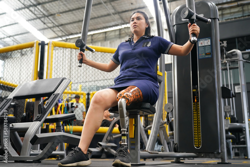 Young female with one prosthetic leg with shoulder exercises with exercise machines to train the muscles in the torso and shoulders, the concept of living a woman's life with a prosthetic leg.