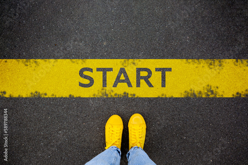 Female feet in yellow sneakers standing on street background with word new start written. Yellow race line beginning idea concept. Top view. Businessman of starting