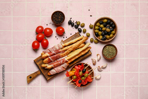 Wooden board of tasty Italian Grissini with bacon  tomatoes and olives on pink tile background