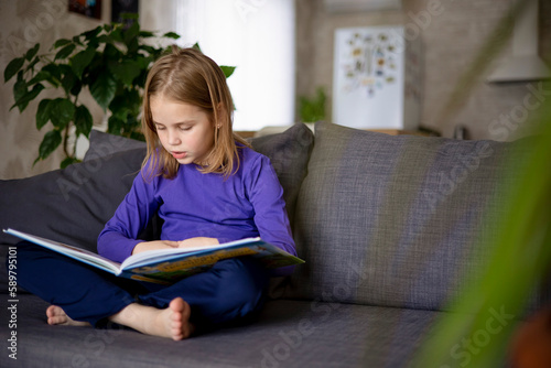 A cute little blonde girl is reading a large colorful book sitting on the sofa in the living room. Self-education of younger schoolchildren, reading literature at home, interesting leisure.