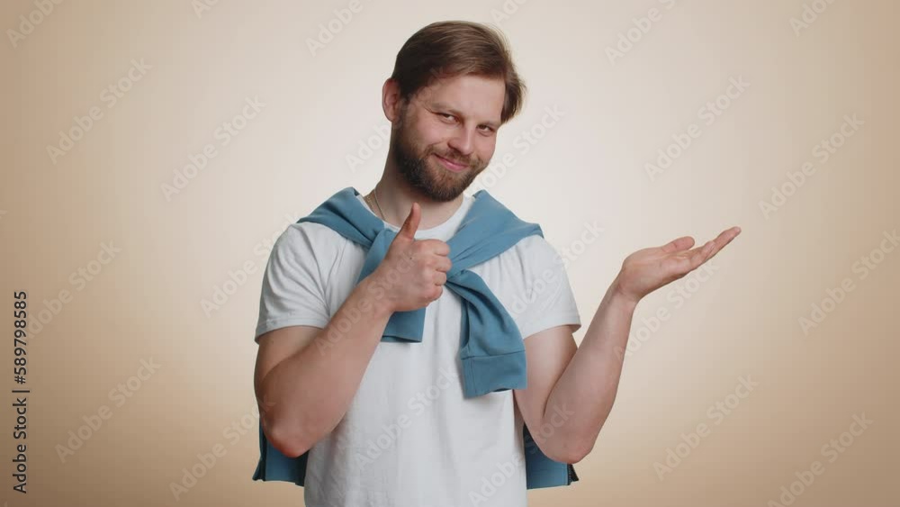Caucasian man showing thumbs up and pointing empty place, advertising area for commercial text, banner, logo, copy space for goods promotion. Young bearded guy isolated on beige studio background Caucasian man showing thumbs up and pointing empty place, advertising area for commercial text, banner, logo, copy space for goods promotion. Young bearded guy isolated on beige studio background