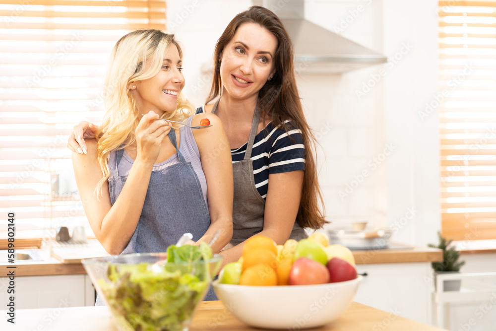 Happy same sex couple feed each other at kitchen. Cheerful LGBTQ lesbian woman feeding salad each other. A beautiful couple of lesbian ladies having a romantic moment in the kitchen.