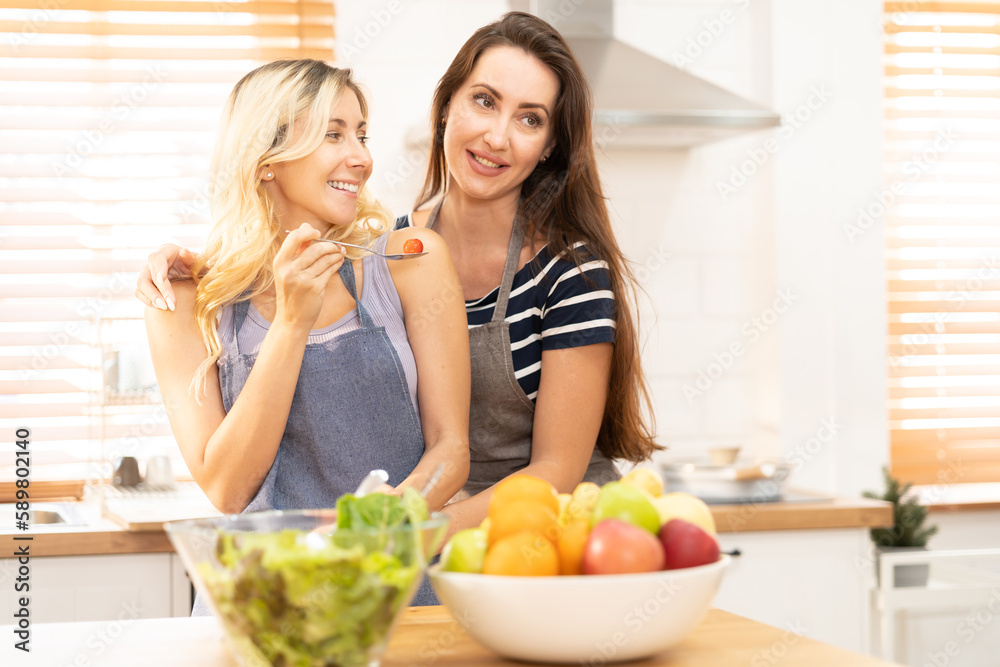 Happy same sex couple feed each other at kitchen. Cheerful LGBTQ lesbian woman feeding salad each other. A beautiful couple of lesbian ladies having a romantic moment in the kitchen. Happy same sex couple feed each other at kitchen. Cheerful LGBTQ lesbian woman feeding salad each other. A beautiful couple of lesbian ladies having a romantic moment in the kitchen.