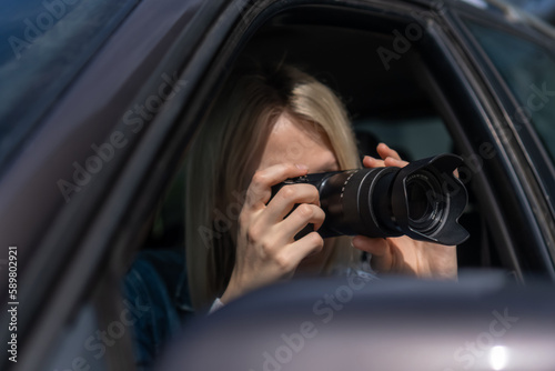 Young blonde woman takes pictures sitting in a car. Concept of journalism, detective, papparation © Volha