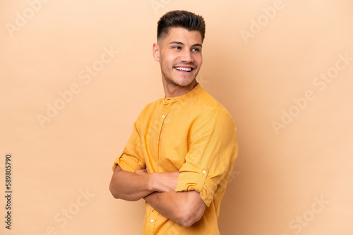 Young caucasian man isolated on beige background looking to the side and smiling