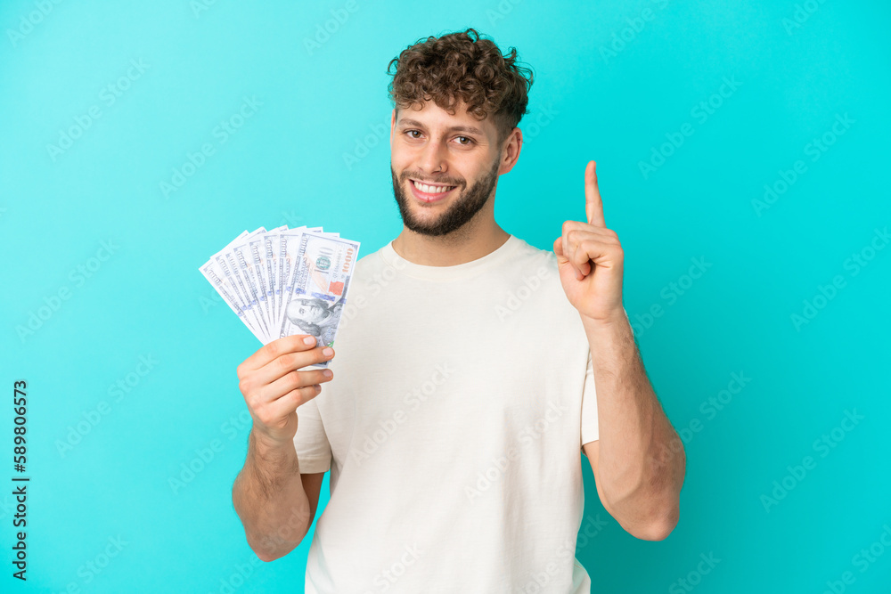 Young caucasian man taking a lot of money isolated on blue background showing and lifting a finger in sign of the best Young caucasian man taking a lot of money isolated on blue background showing and lifting a finger in sign of the best