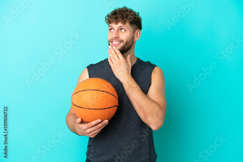 Handsome young man playing basketball isolated on blue background looking up while smiling © luismolinero