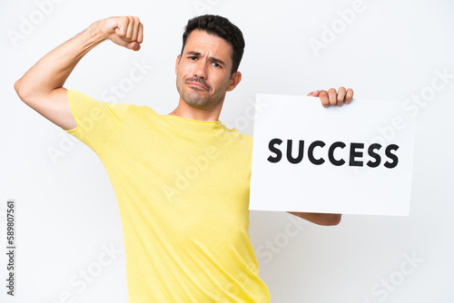 Young handsome man over isolated white background holding a placard with text SUCCESS and doing strong gesture © luismolinero