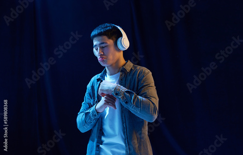 Evaluating songs. Young asian guy in casual stylish clothes listening to music in headphones against dark background in neon light. Concept of human emotions, youth, fashion, lifestyle © master1305