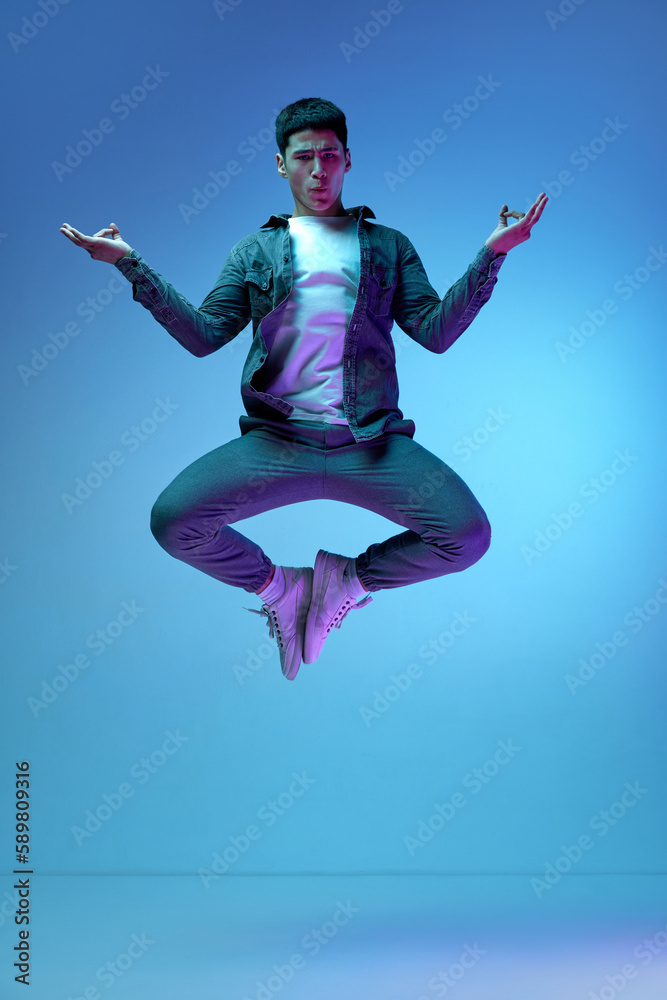 Relaxation, calming down. Young emotive man in casual clothes jumping in yoga pose against blue background in neon light. Concept of human emotions, youth, fashion, lifestyle, feelings