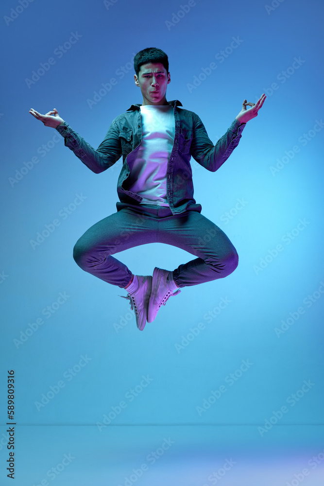 Relaxation, calming down. Young emotive man in casual clothes jumping in yoga pose against blue background in neon light. Concept of human emotions, youth, fashion, lifestyle, feelings Relaxation, calming down. Young emotive man in casual clothes jumping in yoga pose against blue background in neon light. Concept of human emotions, youth, fashion, lifestyle, feelings