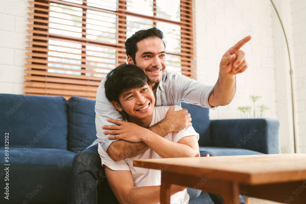 Fototapeta premium Happy caucasian and Asian LGBT couple, one sitting on the sofa embracing the other sitting on the floor from the back at home. Diversity of LGBT relationships. A gay couple concept.