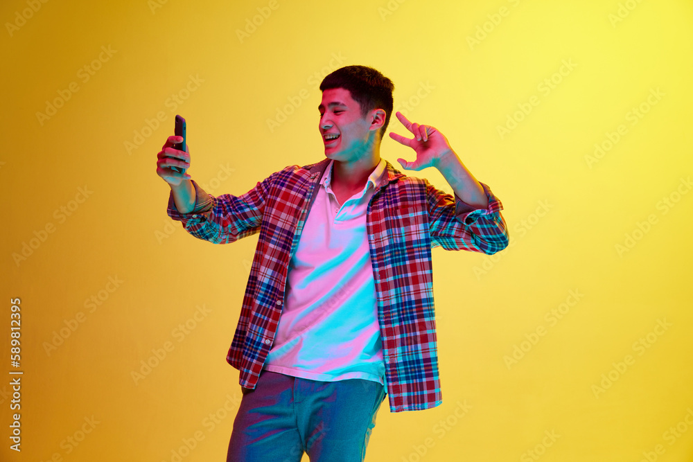 Positive, smiling, young guy taking selfie with mobile phone, posing against yellow background in neon light. Social media influencer. Concept of human emotions, youth, fashion, lifestyle Positive, smiling, young guy taking selfie with mobile phone, posing against yellow background in neon light. Social media influencer. Concept of human emotions, youth, fashion, lifestyle
