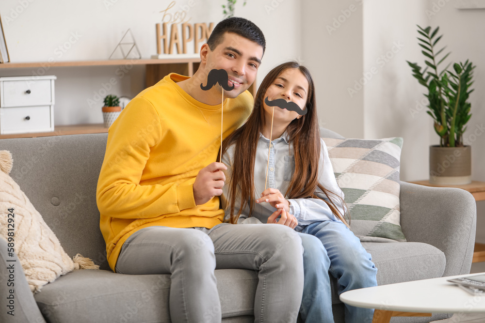 Portrait of father and his little daughter with paper mustache sitting on sofa