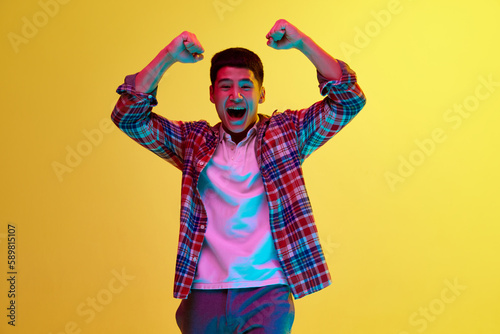 Young korean guy in stylish casual clothes showing emotions of success, win against yellow background in neon light. Achievement. Concept of human emotions, youth, fashion, lifestyle