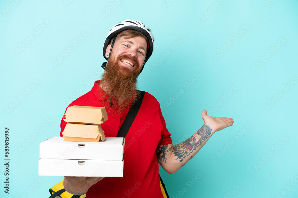 Delivery man holding pizzas and burgers isolated on blue background extending hands to the side for inviting to come Delivery man holding pizzas and burgers isolated on blue background extending hands to the side for inviting to come