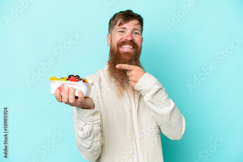 Redhead man with long beard holding a bowl of fruit isolated on blue background and pointing it
