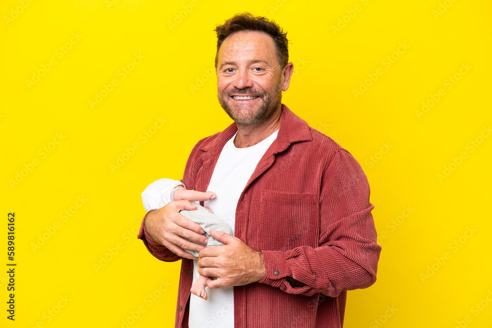 Middle age caucasian man holding his newborn son isolated on yellow background smiling a lot Middle age caucasian man holding his newborn son isolated on yellow background smiling a lot