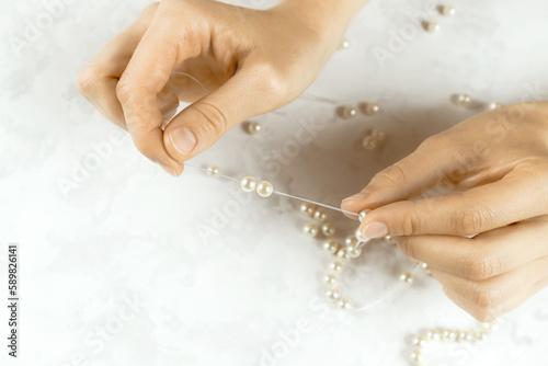 Close-up of woman hands stringing pearls on a necklace on white background. Beads jewellery making process. Hobby handmade concept. Top down view. Selective focus. Blurred background.
