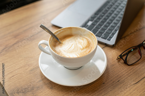 Moment of rest. Cup of fragrant cappuccino coffee with foam is standing on wooden table next to laptop. Coffee break. 