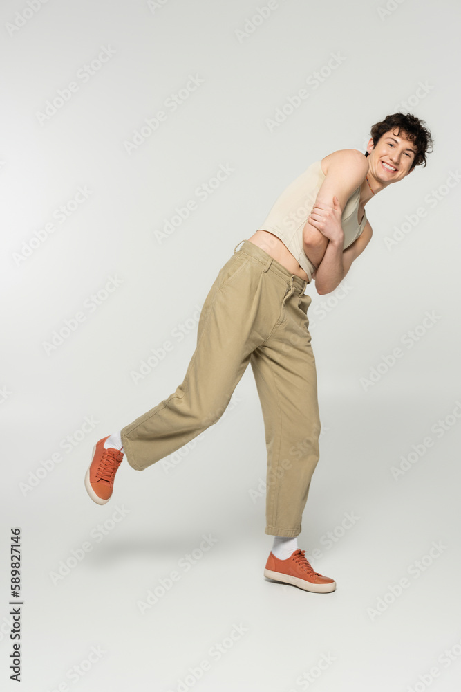 full length of cheerful nonbinary person in beige pants and sneakers posing with folded arms on grey background.